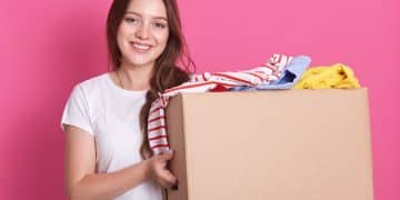 Horizontal shot of female stands smiling near both with reusable clothing for poor peoplw isolated over pink background in studio, attractive woman making charity, posing with happy expression.